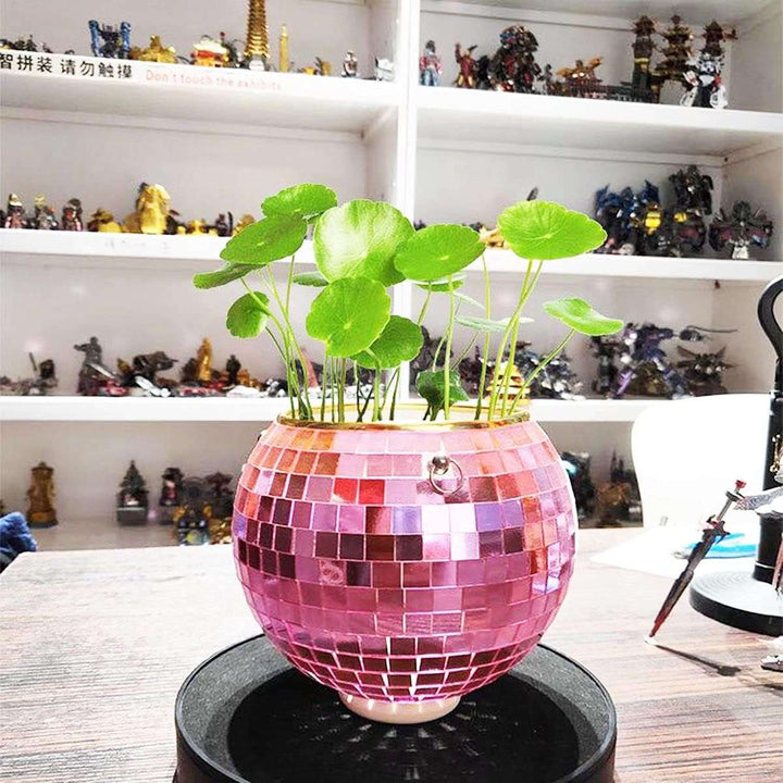 VibeCore Disco Ball Planter in pink, placed on a side table, featuring a shiny, reflective mirror-ball surface. This retro-inspired planter adds a fun and colorful touch to any room while showcasing your plants in style. The pink disco ball design brings a playful, vibrant atmosphere to your living room, bedroom, or office decor.