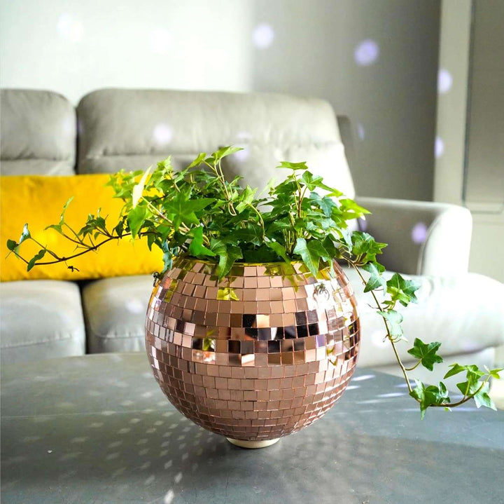 VibeCore Disco Ball Planter in rose pink, placed on a living room coffee table, featuring a shiny, reflective mirror-ball surface. This retro-inspired planter adds a playful pop of color and a fun, vibrant vibe to your space while showcasing your plants. Perfect for enhancing the decor of your living room with its unique and eye-catching design.