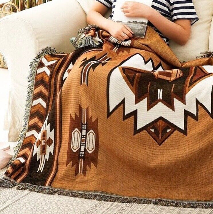 VibeCore Brown Aztec Woven Blanket being used by a person on a couch, featuring a tribal-inspired design, reversible patterns, and soft, breathable 100% combed cotton. Adds cozy, vibrant style and earthy charm to the space.