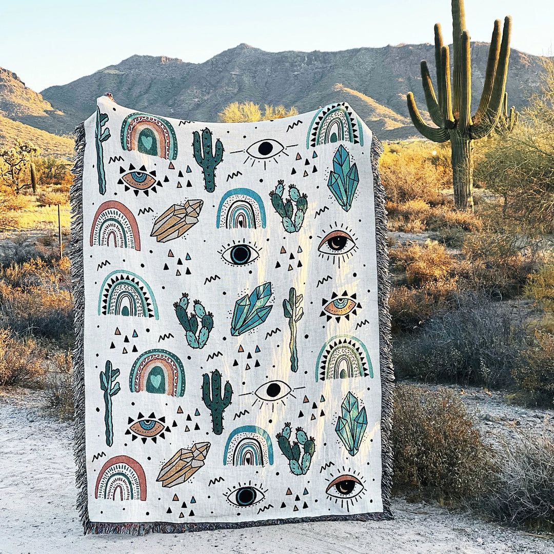 VibeCore Cactus & Crystals Woven Blanket held up in a desert setting, highlighting its earthy cactus and crystal design against a natural landscape, with soft, reversible cotton texture.