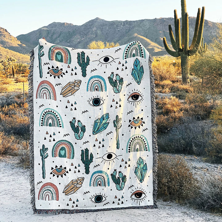 VibeCore Cactus & Crystals Woven Blanket held up in a desert setting, highlighting its earthy cactus and crystal design against a natural landscape, with soft, reversible cotton texture.