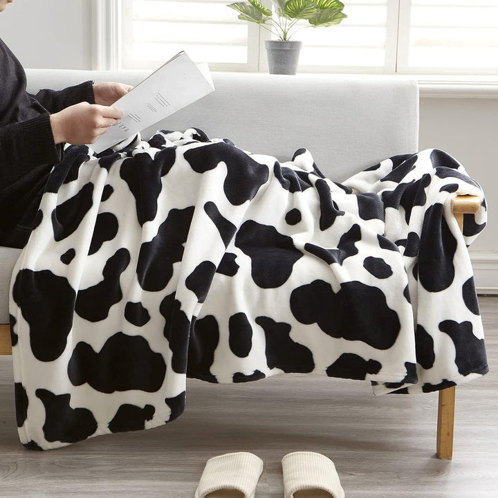 Person cuddled in a black cow print throw blanket, reading a book, showcasing the cozy, playful design and soft texture. Perfect for adding comfort and style to any relaxing moment.