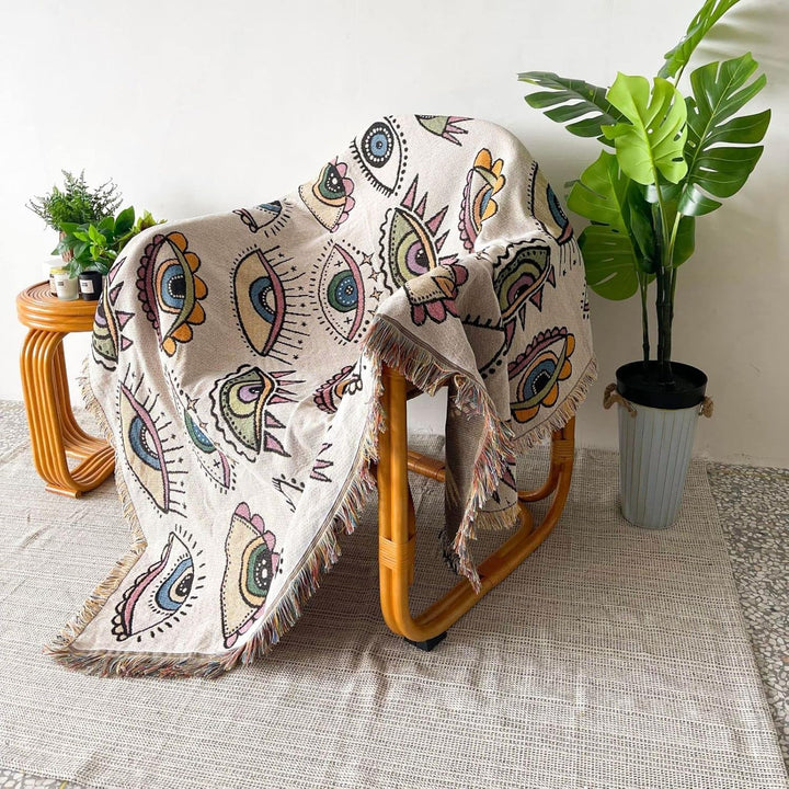 VibeCore Eternal Gaze Woven Blanket on chair, featuring an intriguing and bold design. Made from soft, breathable 100% combed cotton, it adds vibrant energy, mystique, and artistic flair to your space.