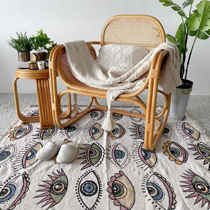 VibeCore Eternal Gaze Woven Blanket on floor under chair, featuring a captivating and bold design. Made from soft, breathable 100% combed cotton, it adds vibrant energy and mystique while enhancing the cozy atmosphere of your space.
