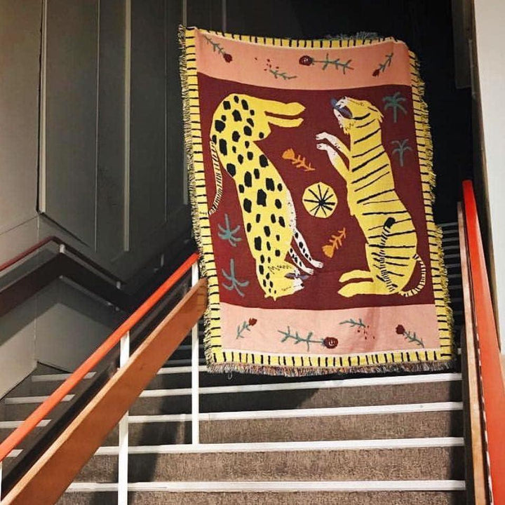 VibeCore Feline Power Woven Blanket held up on staircase, featuring a bold jungle design in soft 100% cotton for a striking and cozy addition to your space.