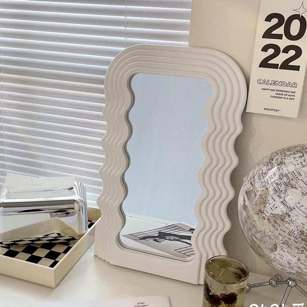 VibeCore Retro Wave Mirror in white, leaning against a wall and placed on a desk, with a wave-shaped resin frame and sleek mirror lens, adding a modern, eclectic touch to the workspace.