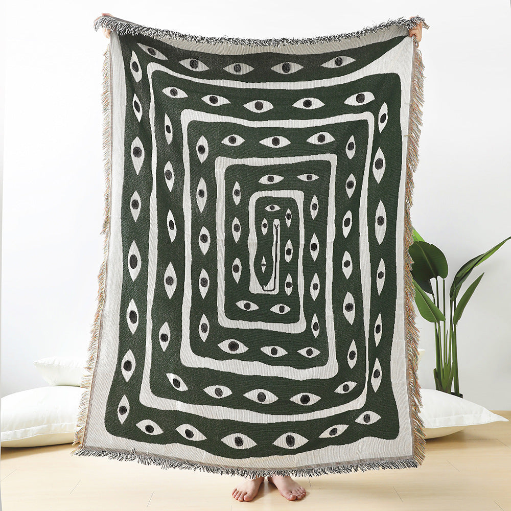 VibeCore Snake-Eye Woven Blanket held up in a front view, highlighting its bold snake-eye design. Crafted from 100% combed cotton, this versatile and striking piece adds a touch of mysterious elegance to any space.
