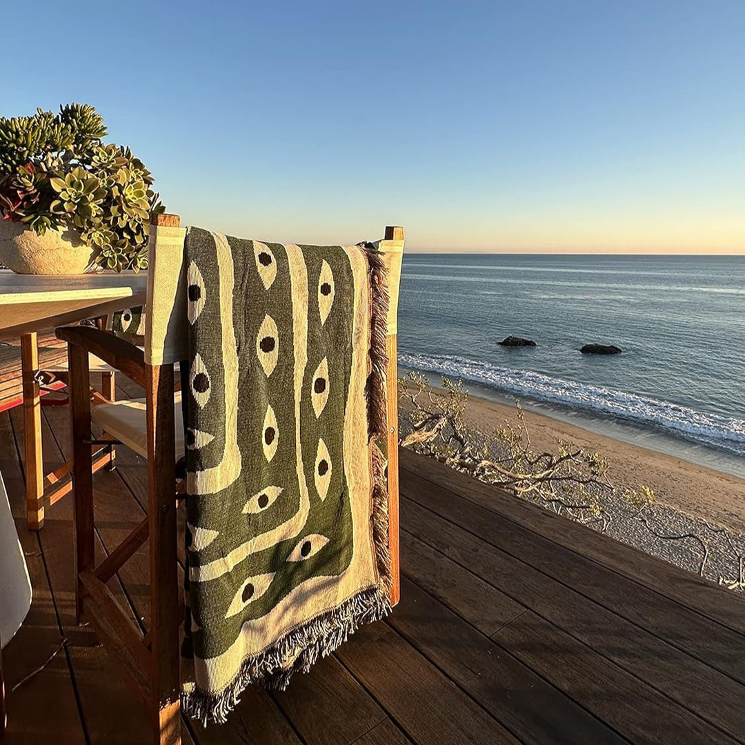 VibeCore Snake-Eye Woven Blanket draped over a chair by the ocean, featuring its bold snake-eye design. Made from 100% combed cotton, this versatile blanket adds a touch of mystery and elegance to your space while complementing coastal vibes.