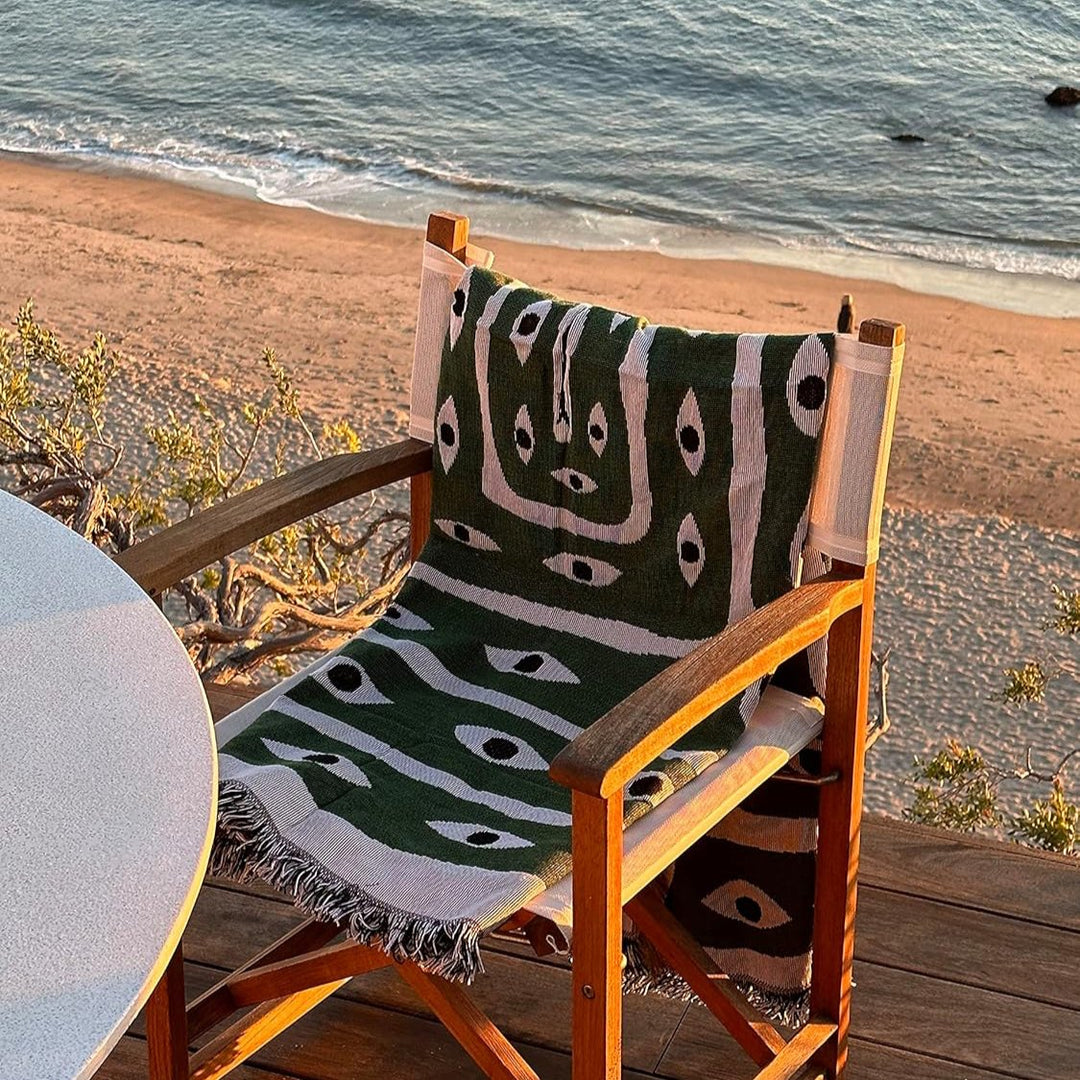 VibeCore Snake-Eye Woven Blanket draped over a chair by the ocean, captured from an alternative view. The bold snake-eye design, made from 100% combed cotton, combines edgy style and coastal serenity, bringing a unique, mysterious touch to your surroundings.
