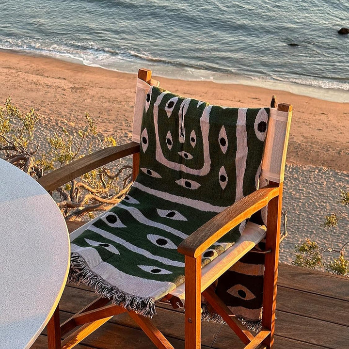 VibeCore Snake-Eye Woven Blanket draped over a chair by the ocean, captured from an alternative view. The bold snake-eye design, made from 100% combed cotton, combines edgy style and coastal serenity, bringing a unique, mysterious touch to your surroundings.