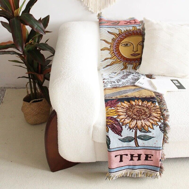 VibeCore The Sun Tarot Woven Blanket folded on couch, featuring a celestial-inspired design with vibrant, reversible patterns. Made from soft, breathable 100% combed cotton, it adds a cosmic touch to your space while offering versatile styling options with its folded, neatly arranged look.