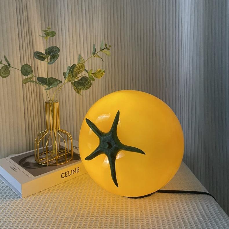 A glowing yellow glass tomato-shaped table lamp, radiating a soft and inviting light. Perfect for adding a quirky yet cozy touch to eclectic home decor.