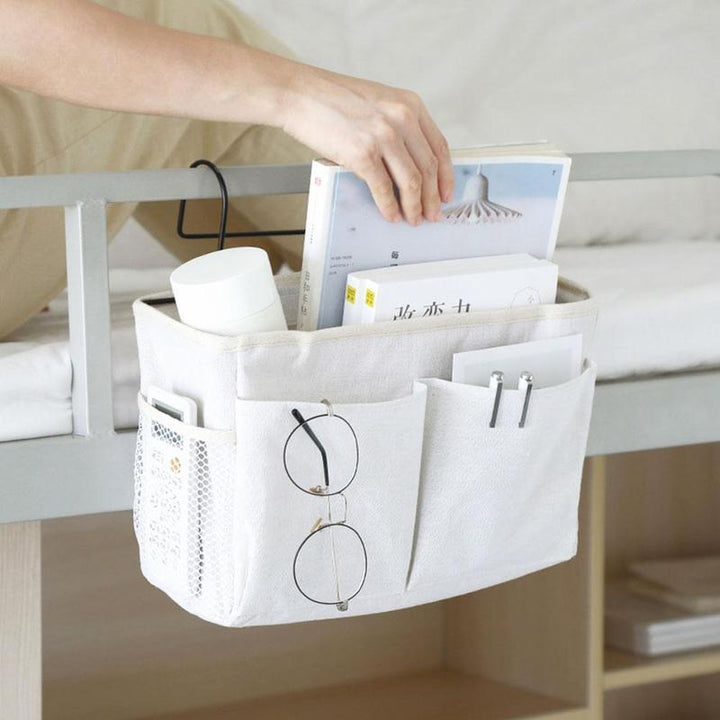 White VibeCore bedside caddy placed beside a bed, featuring multiple pockets and hooks for storing essentials like phones, tablets, and books. Durable, portable, and space-saving, ideal for small spaces like dorms and apartments.